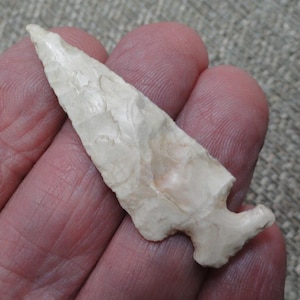May include: A cream-colored stone arrowhead featuring a sharp, pointed tip and a notched base. The surface displays a textured, flaked appearance with subtle variations in tone, showcasing a pointed triangular shape with a distinct stem.