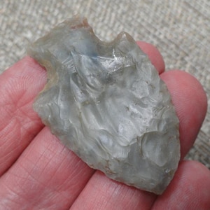 May include: A translucent, light gray stone arrowhead featuring a chipped, textured surface with visible flaking patterns. The object has a pointed tip and a notched base, showcasing a smooth, glass-like appearance across its irregular, sharp edges.