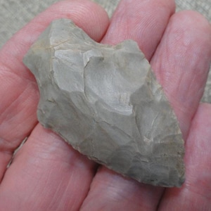May include: A light gray, triangular stone arrowhead featuring a chipped, textured surface with visible flaking patterns along the edges. The object has a pointed tip and a wider, notched base, displaying a matte finish and a solid, opaque appearance.