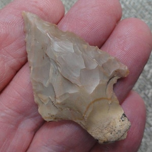 May include: A light tan, triangular stone arrowhead featuring sharp, serrated edges and a pointed tip. The surface displays distinct, layered flake patterns created by knapping, with a rough, unworked base section at the bottom of the object.