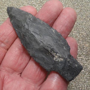 May include: A dark gray arrowhead with a sharp, pointed tip. The stone artifact has a textured surface and a roughly rectangular base. The arrowhead is held in a hand, showcasing its size and detail.