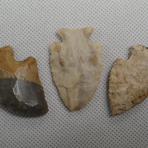 May include: Three stone arrowheads arranged side by side. The left arrowhead is tan and dark gray with a notched base. The center arrowhead is pale cream with a pointed tip and notched base. The right arrowhead is light tan with a textured surface.
