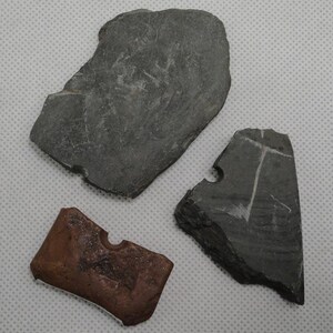 May include: Three irregular stone fragments consisting of two dark gray pieces with rough, uneven edges and one smaller, rectangular reddish-brown piece. Each fragment features a small, semi-circular notch carved into one of its straight edges.