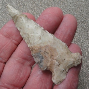 May include: A triangular stone arrowhead featuring a pointed tip and a notched base. The surface is textured with visible knapping marks and displays a color palette of off-white, cream, and light brown with a small patch of dark gray stone.