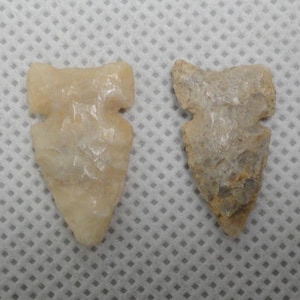 May include: Two stone arrowheads. One is a light beige color, and the other is a mix of beige and gray. Both have a triangular shape with a notched base. The arrowheads are likely handmade and show signs of wear.
