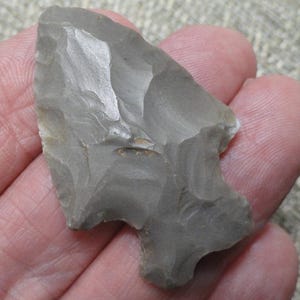 May include: A gray stone arrowhead featuring a pointed tip and a notched base. The surface of the object displays distinct flaked edges and a smooth, matte texture characteristic of knapped stone, showing a triangular shape with defined side notches.