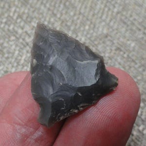 May include: A triangular-shaped arrowhead crafted from dark gray flint stone. The surface features distinct, sharp-edged flake patterns created through knapping, resulting in a pointed tip and a wider, slightly concave base edge.