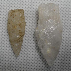 May include: Two stone arrowheads with pointed tips and jagged edges. The smaller arrowhead on the left is tan with brown mineral inclusions, while the larger arrowhead on the right is translucent white quartz with a crystalline, textured surface.