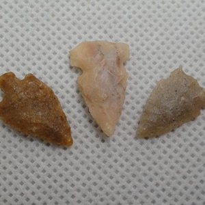 May include: Three antique arrowheads in shades of brown and beige are displayed against a white patterned background. The arrowheads vary in shape and size, showcasing the craftsmanship of ancient tools.