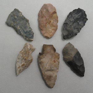 May include: A collection of six stone arrowheads arranged in two rows. The pieces feature varied textures and colors, including shades of gray, tan, brown, and reddish-pink. Each arrowhead displays distinct flaked edges and pointed tips.
