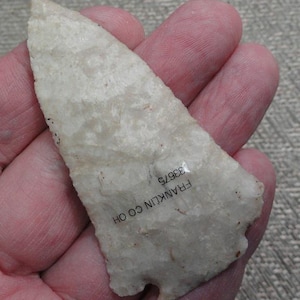 May include: A light-colored, chipped stone arrowhead with a triangular shape. The arrowhead is held in a hand and has the text "FRANKLIN CO OH" and "33675" printed on it.