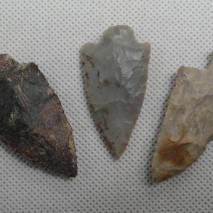 May include: Three stone arrowheads with pointed tips and notched bases. The left arrowhead is dark brown and textured, the center one is smooth light gray, and the right one is a mottled tan and beige color with visible flaked edges.