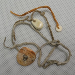 May include: A collection of handmade shell and bone pendants on knotted twine. The pendants are in shades of cream, tan, and brown. The twine is a light beige color, and the overall aesthetic is rustic and natural.