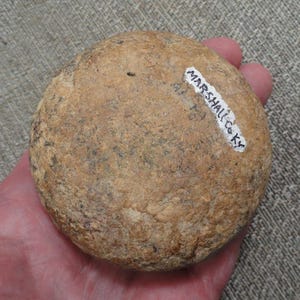 May include: A round, tan-colored stone artifact with a rough, textured surface. A white adhesive label is attached to the top right side, featuring the black handwritten text MARSHALL CO KY. The object has a circular shape and earthy tones.
