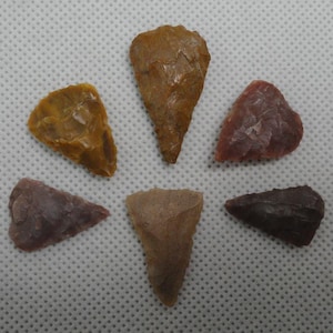 May include: Six antique arrowheads in shades of brown, tan, and reddish-purple are arranged in a circular pattern. Each arrowhead has a distinct shape and texture, showcasing the craftsmanship of a bygone era. The background is a white, textured surface.