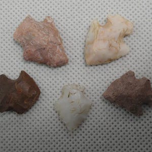 May include: Five ancient arrowheads in varying shades of brown, pink, and white are displayed. Each arrowhead has a unique shape and texture, showcasing the craftsmanship of a bygone era. The arrowheads are arranged on a textured, light-colored surface.