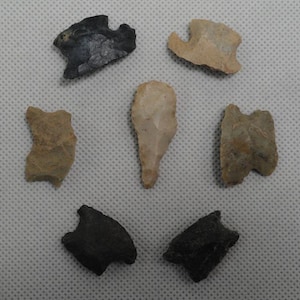 May include: A collection of six stone arrowheads arranged in a circular pattern. The set includes two dark charcoal-colored arrowheads, three tan-colored arrowheads with textured surfaces, and one central elongated light tan stone point.