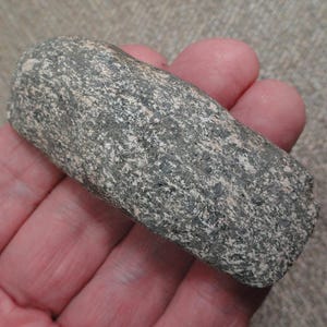 May include: A rectangular stone tool with a rough, speckled texture featuring a mix of dark gray, black, and light tan mineral grains. The object has rounded edges and a weathered, natural appearance with a dense, granular surface composition.
