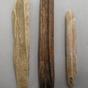 May include: Three weathered wooden artifacts of varying sizes and shades of brown and tan. The central piece is a dark brown, rectangular shape with a carved groove. The other two are lighter in color, with one having a similar groove.