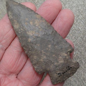 May include: A gray stone arrowhead with a pointed tip and a notched base. The arrowhead has a rough, textured surface with areas of lighter brown coloration. The text "GRAVES CO. KY." is visible on the base.