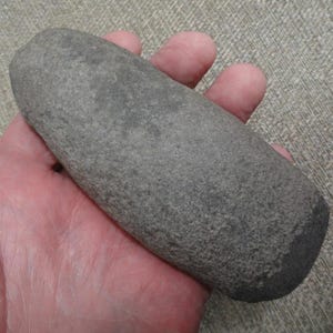 May include: A gray, oblong stone, possibly a grinding tool, held in a hand. The stone has a rounded end and a textured surface. The color is a muted gray, with darker areas suggesting wear or age. The object is a natural, hand-held item.