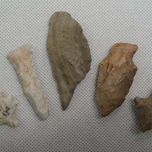 May include: Five ancient stone tools, including arrowheads and blades, are displayed. The artifacts vary in color from light beige to brown, with visible chipping and wear. One arrowhead has the markings "LAS" on it.