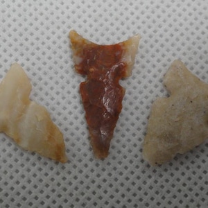 May include: Three ancient arrowheads in shades of tan and brown are displayed against a white patterned background. The central arrowhead is a deep reddish-brown, while the others are lighter in color, showcasing the natural variations in the stone.