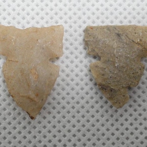 May include: Two ancient arrowhead artifacts. One is a light tan color, the other is a darker tan with a crack. Both have a distinctive notched design, likely used for hunting or warfare.