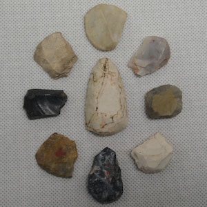 May include: A collection of nine assorted stone artifacts arranged in a circular pattern. The pieces vary in color, including shades of white, tan, gray, brown, and black, with irregular, chipped edges and textured surfaces characteristic of flint knapping.