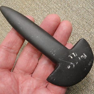 May include: A dark gray, smooth, T-shaped stone tool with a rounded, elongated handle and a wide, curved base. The surface features white handwritten text that reads "Port Co ILL" on the flat, curved section of the stone artifact.