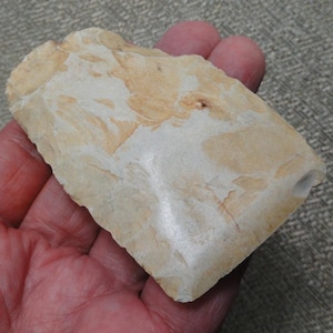 May include: A hand-knapped stone tool featuring a flat, wedge-shaped profile with a smooth, polished edge. The surface displays a marbled pattern of cream, beige, and light tan colors with visible flake scars across the textured stone body.