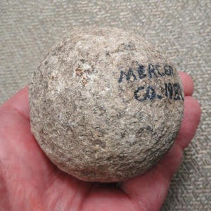 May include: A spherical, textured gray stone artifact with a rough, weathered surface. The object features black handwritten text on its side that reads MERCER CO. 1981, indicating its origin and date. The stone has a mottled appearance.