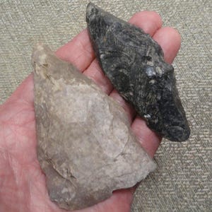May include: Two ancient stone tools, one light gray and the other dark gray, are held in a hand. The tools have sharp edges and a rough texture, suggesting they were used for cutting or scraping. The background is a neutral, textured surface.