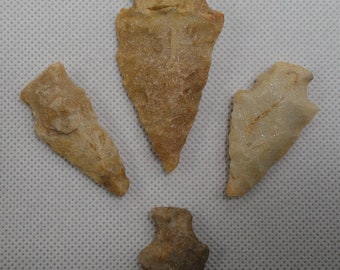 May include: Four ancient stone arrowheads in varying shapes and sizes. The arrowheads are primarily beige and tan, with visible textures and edges. The arrangement suggests a collection of historical artifacts, possibly for display or study.