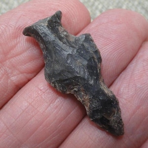 May include: A dark gray, chipped stone arrowhead featuring a pointed tip, a central ridge, and a notched base. The surface displays a textured, flaked appearance characteristic of knapped stone, with variations in color from charcoal to black.