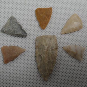 May include: A collection of six ancient arrowheads in various shapes and colors. The arrowheads are made of stone and range in color from gray to tan and orange. The arrowheads are arranged on a white surface.