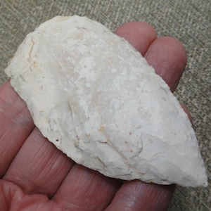May include: A pale, off-white stone artifact with a pointed, leaf-shaped design and chipped, textured edges. The surface displays a matte finish with subtle variations in tone, featuring a sharp, tapered tip and a rounded, wider base.