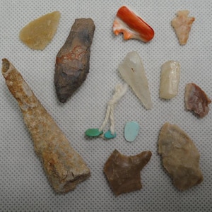 May include: A collection of various small, ancient artifacts, including arrowheads, bone fragments, and polished stones. The items are in shades of brown, beige, and orange, with a few turquoise and white accents. The objects are arranged on a white, textured surface.