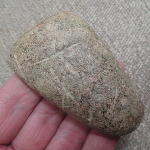 May include: A hand-held, oblong stone tool with a rounded end and a cross-like etched design. The stone is a mottled gray-brown color, with a rough, textured surface. The tool appears to be an ancient artifact.