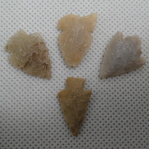 May include: Four ancient arrowheads in varying shades of tan and beige are displayed against a white background. The arrowheads have a triangular shape with a notched base, showcasing their historical design.