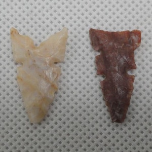 May include: Two stone arrowheads. One is a light beige color, and the other is a deep reddish-brown. Both arrowheads have a similar shape, with a notched base and a pointed tip. The arrowheads are likely of Native American origin.