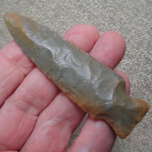 May include: A pointed stone projectile point featuring a flaked surface texture with a color palette of muted gray and earthy brown tones. The object has a tapered blade shape with a notched base, showcasing distinct knapped edges throughout.