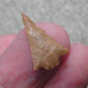 May include: A tan-colored arrowhead with a triangular shape and a sharp point. The arrowhead is held between two fingers, showcasing its texture and craftsmanship. The stone tool appears to be an ancient artifact.
