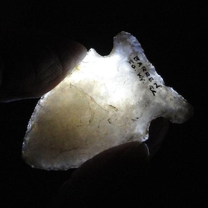 May include: A translucent, off-white arrowhead with a notched design. The arrowhead has a pointed tip and a curved base. The text "BARREN CO. KY." is written on the surface. The arrowhead is held against a dark background.