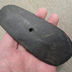 May include: A dark gray, oblong stone object with a rounded end and a hole near the center. The surface has a slightly rough texture and some visible scratches. The text "701 FICKRA CO." is etched into the stone. The object is held in a hand.