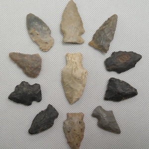 May include: A collection of ten ancient arrowheads, arranged in a circular pattern. The arrowheads vary in size, shape, and color, ranging from light tan to dark gray. They are made of stone and show signs of wear and age.