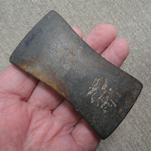 May include: A rectangular, dark gray stone tool with a weathered, textured surface. The object features visible scratches, patches of brown discoloration, and the handwritten text "L-V-II" in black ink on one of the beveled edges.