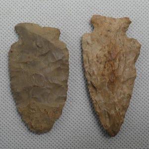 May include: Two stone arrowheads with chipped, textured surfaces. The left arrowhead is a muted grayish-brown with a rounded base, while the right arrowhead is a lighter tan color with a more pointed tip and distinct notched edges.