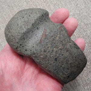 May include: A gray stone artifact with a rounded head and a rectangular base. The stone has a rough, textured surface with small dark flecks and a reddish-brown streak. The object is held in a hand.