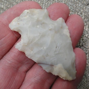 May include: A white, translucent stone arrowhead featuring a triangular shape with notched sides and a pointed tip. The surface displays a textured, flaked appearance with subtle variations in opacity and small, natural inclusions throughout.